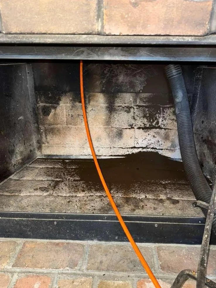 A soot-stained brick firebox with an orange cable running down its middle and a ribbed black vacuum hose at the right, under a dark steel lintel