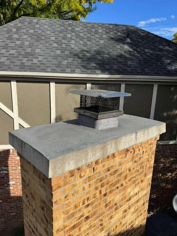 An orange-brown brick chimney with a flat grey concrete top and a steel cap with black mesh, in front of a grey hip roof and a tan and brown wall