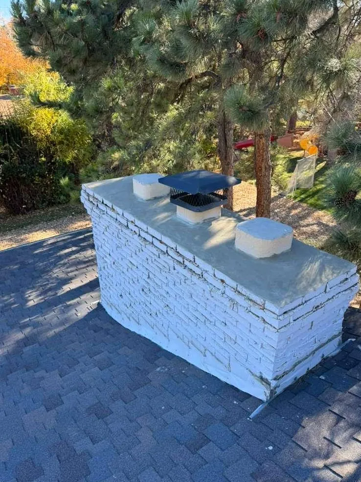 A white-painted brick chimney seen from above, with a blue steel cap and two pale clay flue tops on a concrete crown, among ponderosa pines and autumn leaves