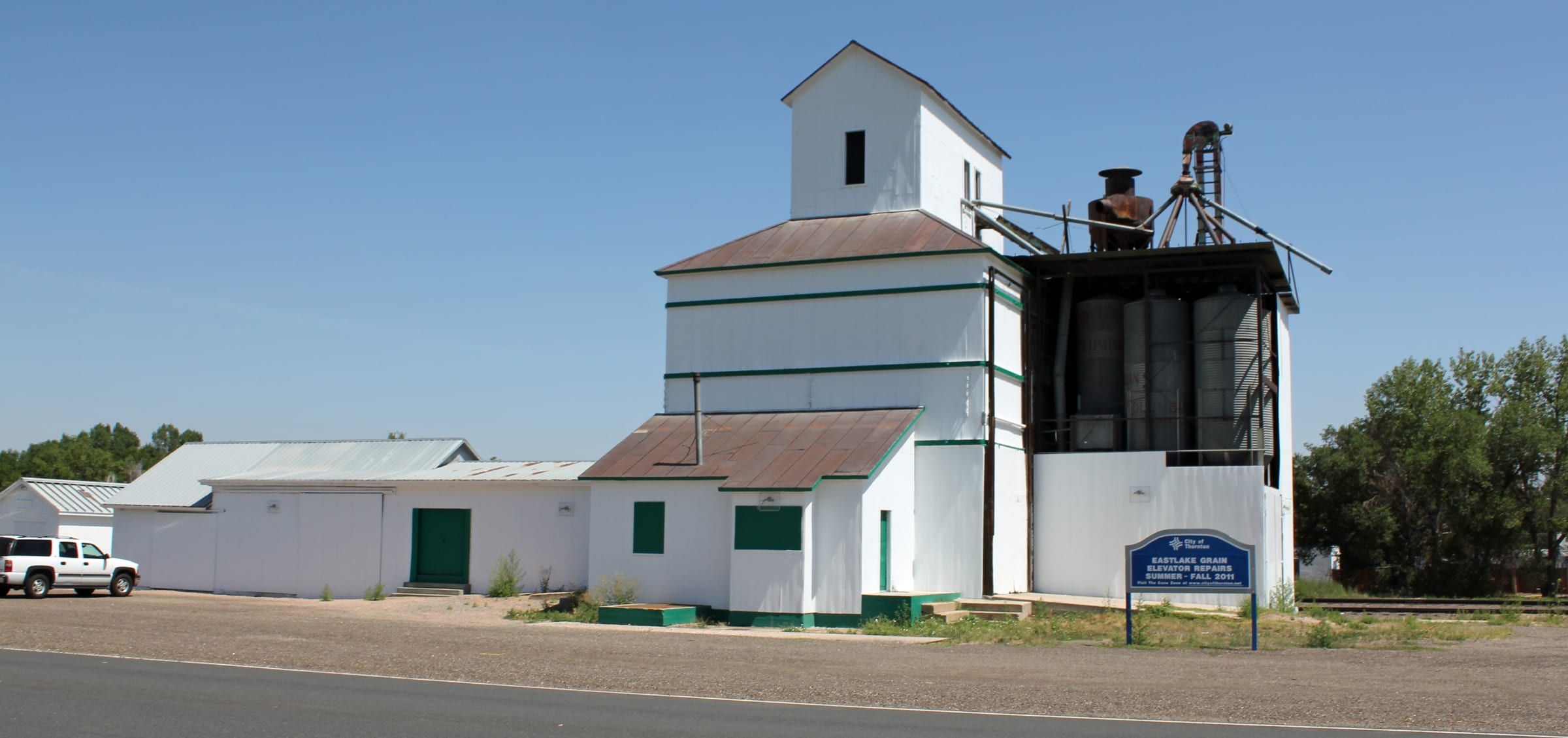 A white-painted timber grain elevator with green trim and rust-red roofs beside a gravel lot, under a clear blue sky
