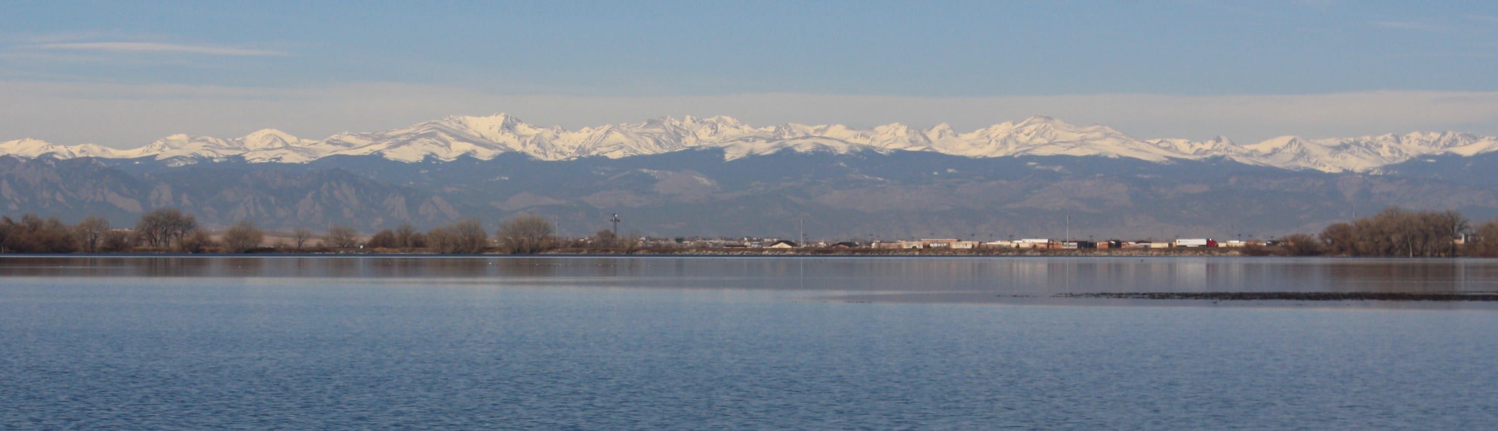 The snow-capped Front Range above a calm lake, with a line of bare trees and low rooftops along the far shore under a pale blue sky